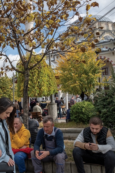 Picture of two women walking in front a many men sitting on a concrete ledge in a crowded park.