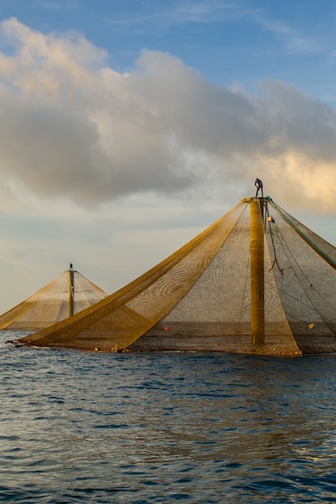 A man seated in a small rowboat extends his arms to catch an object. Behind him, two diamond-shaped fish cages rise from the water.