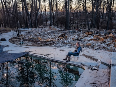 a woman sits in a garden chair around the charred ruins of her home